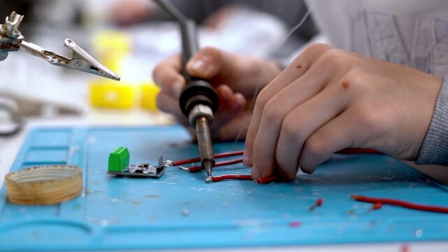 Hands hold soldering iron over circuit board. Red wires lie nearby on blue mat. Focus on precise, careful soldering task. Detail shows concentration and technical skill. Great for electronics, DIY