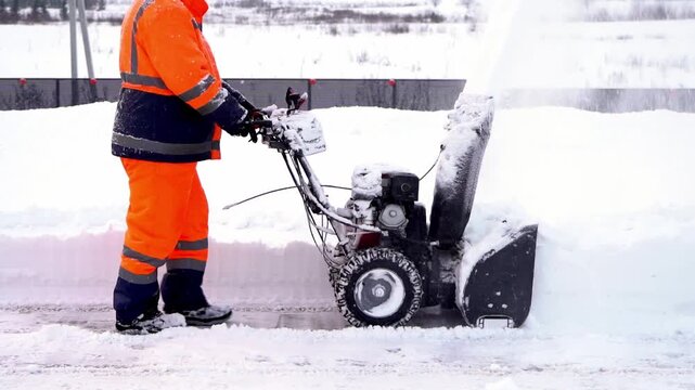 Worker clears snow with motorized snow blower. Snow flies as machine grinds through thick accumulation. Bright orange suit contrasts against white landscape