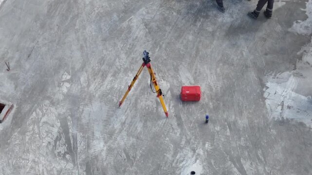 Surveyor's tripod rests on concrete ground. Red case sits nearby, tools nearby. Worker's feet appear in frame. No people express emotion. Ideal for construction or engineering scenes