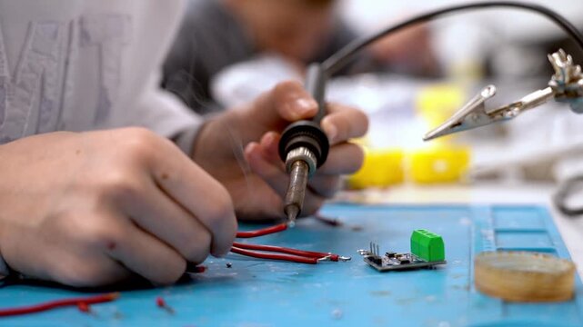 Hands hold soldering iron. Wires lie nearby, awaiting connection. Blue mat holds small electronic parts. Focus is sharp on hands and tools. No facial expressions visible