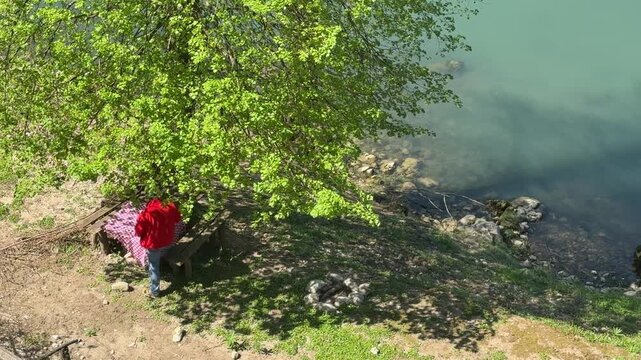 Man sitting at a rustic picnic table under a tree near a river in the countryside. Peaceful outdoor scene with natural shade, simple food, and slow living atmosphere. Ideal for concepts of relaxation