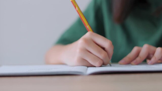 Close up selective focus of a student's hand holding a pencil, writing in a notebook. Bright daylight for the workspace, representing academic concentration,homework, and childhood education concepts.