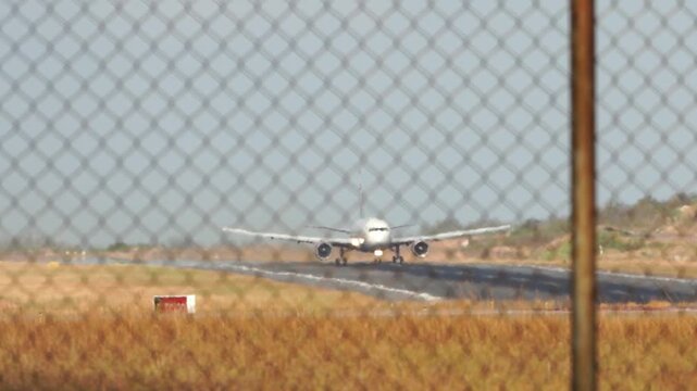 Plane lands on runway behind fence. Chain-link barrier blocks view from foreground. Sunlit desert surrounds airport perimeter. No people visible. Scene captures quiet airport arrival moment