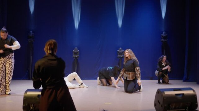Theatre rehearsal on dimly lit stage with director guiding performers during contemporary dance practice in empty auditorium before live performance