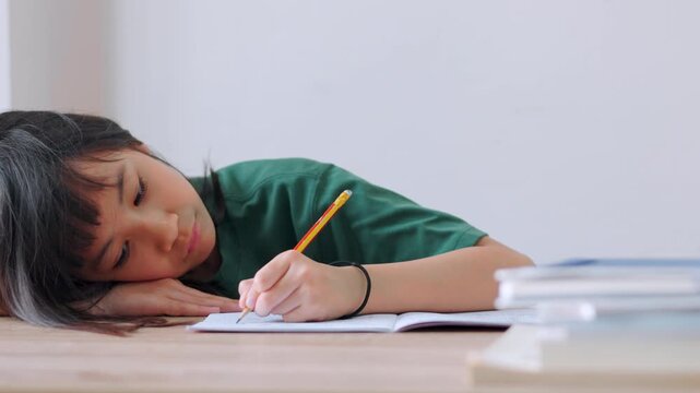 Close up selective focus of a tired Asian kid girl resting her head on the desk. Stressed and exhausted from studying too much homework in bright daylight. Education burnout and overwork concept.