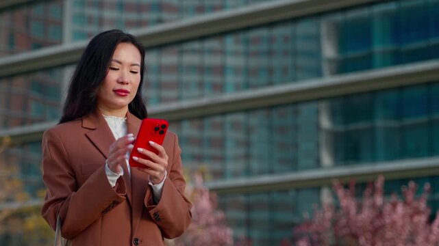 Asian businesswoman using smartphone cellphone mobile phone device outdoors in modern city, business office building background. Woman holding telephone, thoughtful focused expression, copy space