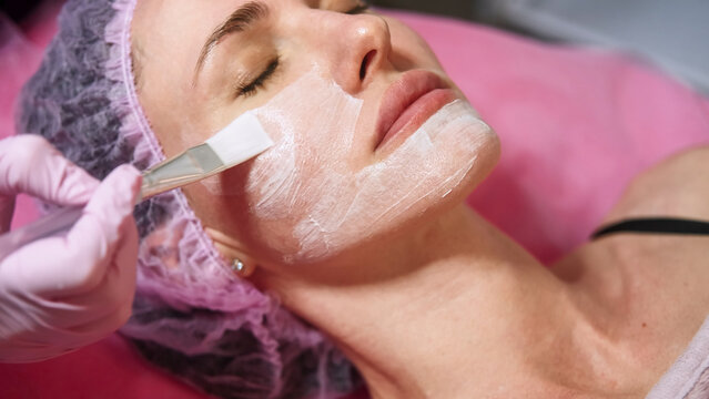 Peeling mask. Skin rejuvenation. Professional beautician hand using brush applying moisturizing cream on female client face for skincare detox procedure at beauty salon.