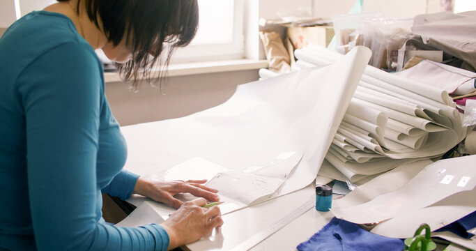 Dressmaking process. Tailor desk. Woman hand creating garment patterns with ruler making notes on table cluttered with paper drafts.