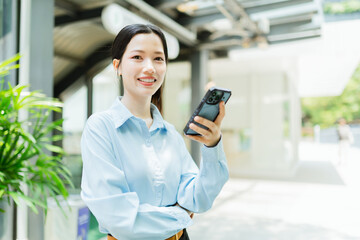 Fototapeta premium Young woman in a blue shirt smiling and holding a smartphone outdoors in a modern urban setting.