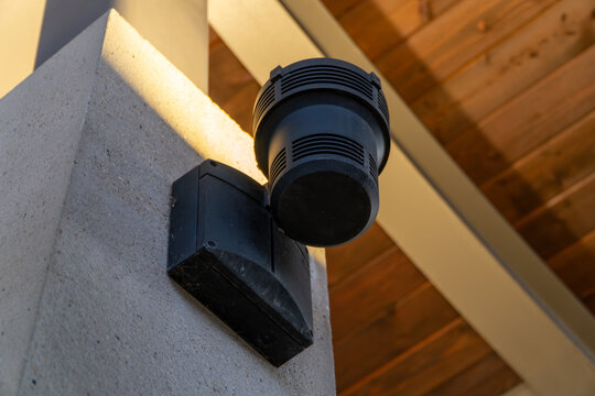 Outdoor black sound system speaker or security siren mounted on textured concrete pillar under wooden ceiling with golden hour sunlight.