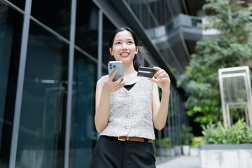 Fototapeta premium A young woman outdoors smiles while holding a smartphone and a credit card near a modern glass building.