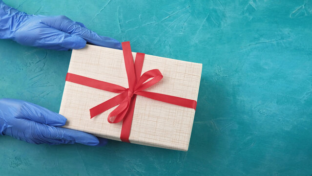 Quarantine surprise. Pandemic birthday. Hands in gloves holding wrapped gift box with red ribbon bow on blue textured background with free space.