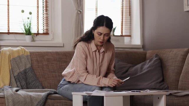 Poverty and debt. Angry young Caucasian woman is sitting on sofa, counting money, checking receipt and throwing away cash. Concept of domestic accounting and utility bills