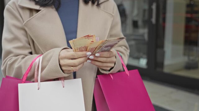 Woman counting pesos with hands while holding pink shopping bags in front of a shop on street, beige coat visible; shopping satisfaction.