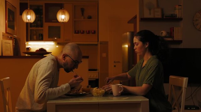 Cozy evening. Young Caucasian couple is having dinner together in kitchen. Bearded bald man and pretty woman eat homemade food at home. Husband and wife date. Valentine's Day Concept
