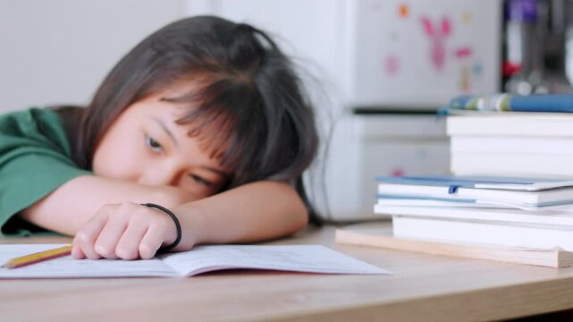 Close up selective focus of demotivated Asian kid girl resting her head on a desk in daylight. She looks overburdened by too much homework, illustrating academic pressure and childhood study burnout.