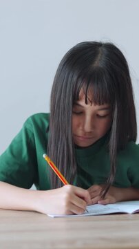 Vertical video showing close up selective focus of a focused asian kid girl writing in a notebook in daylight. Ideal for mobile social media, representing student doing homework and modern education.