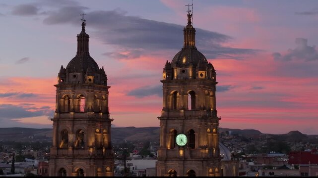 Cathedral Basilica of Immaculate Conception in Durango Mexico towers illuminated against a vibrant pink and orange sunset sky. Colonial baroque architecture with clock tower and crosses over.