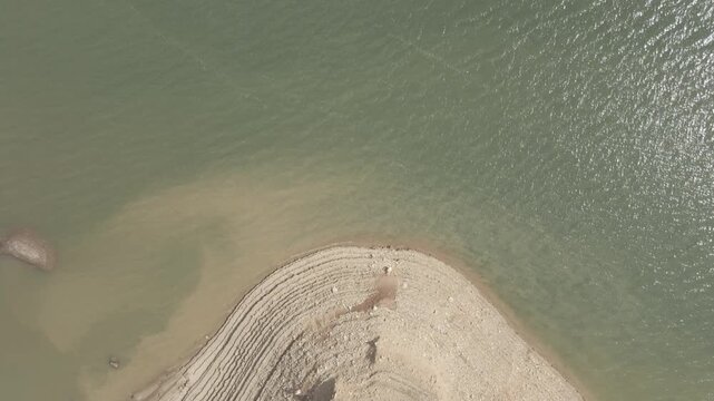 An aerial drone shot begins looking down at water then tilts up revealing a sprawling mountain reservoir experiencing severe drought with low water levels and exposed dry shores.

