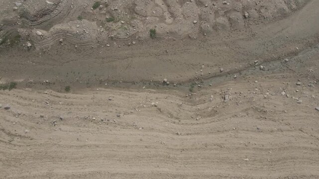 An aerial top down view of a completely dried up riverbed showing layered sandy terrain and scattered rocks, highlighting the severe impact of drought on the natural landscape.

