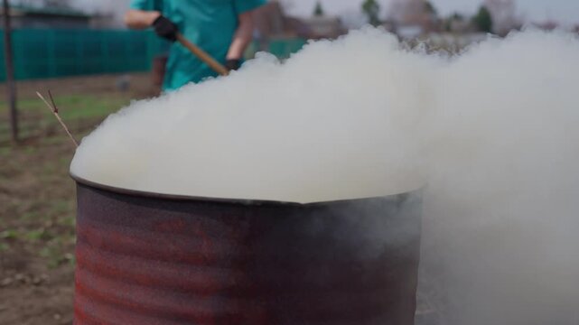 Groundskeeper Raking Leaves Into Smoking Barrel, Thick White Smoke Billowing With Small Flames At Side, Protective Gloves And Quick Motions, Rural Yard Cleanup, Hazard Atmosphere, Ember And Ash