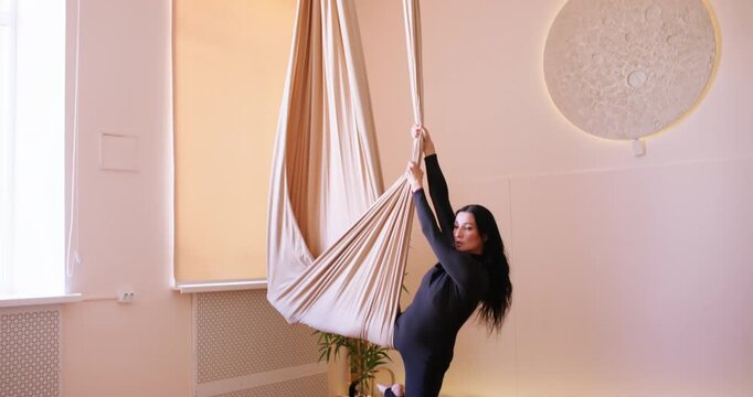 Woman fitness. Muscle strength. Athlete descending from hammock unrolling safety fabric support doing fly yoga side split pose in studio.