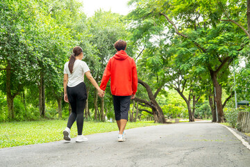 Fototapeta premium Back of Asian man and woman hold hands and walk together along the road in public park with day light for exercise and good activity for better relationship and healthy.
