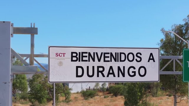 Welcome to Durango Mexico road sign displayed on metal structure along highway. Clear blue sky and green trees frame the official government signage marking the state border entrance.