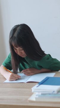 Vertical video showing close up selective focus of a focused asian kid girl writing in a notebook in daylight. Ideal for mobile social media, representing student doing homework and modern education.