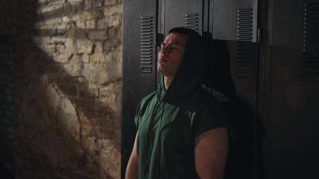 Boxer in green hoodie covering face with bandaged hands in locker room. Male athlete concentrating and breathing before fight in dark gym