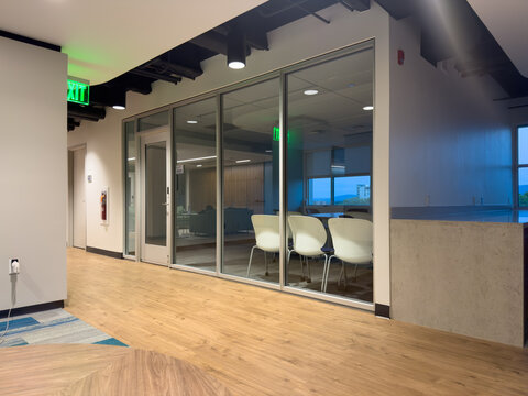 Modern corporate office hallway interior with sleek glass partition walls, exposed industrial ceiling, and warm wood flooring leading to contemporary meeting rooms and a lounge area, featuring a green
