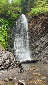 A breathtaking vertical shot capturing a powerful waterfall cascading down a steep, layered rock cliff in a dense green forest.