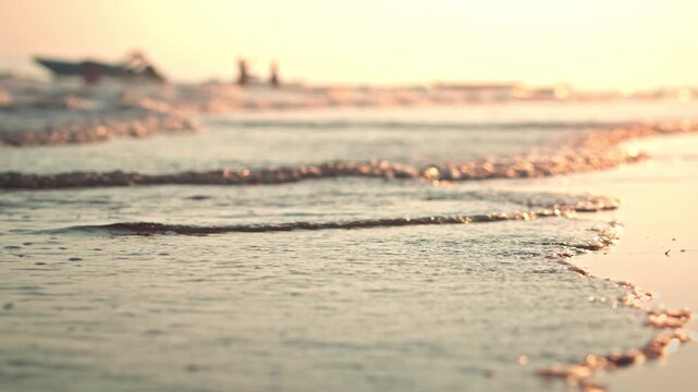 Close-up of gentle sea waves washing onto a sandy beach during golden hour sunset. Beautiful low-angle view with shallow depth of field, Peaceful mood of a summer vacation at the tropical shore.