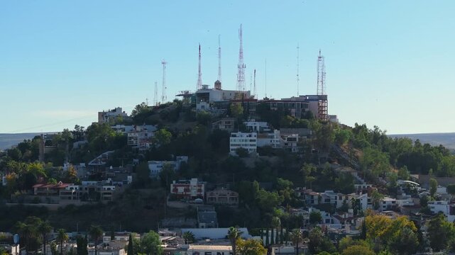 Panoramic view of communication towers crowning a hill surrounded by residential buildings in Durango Mexico. White houses and green trees cover the slopes under clear blue sky.