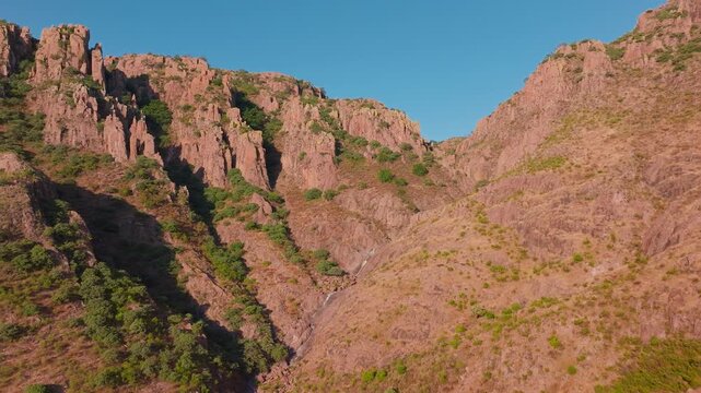 Aerial drone footage captures stunning red rock canyon with dramatic cliff formations and green vegetation dotting steep mountainsides under clear blue sky in rugged Sierra Madre wilderness terrain.
