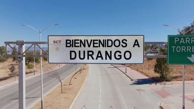 Aerial drone footage of welcome sign at highway entrance to Durango, Mexico. White road sign reads Bienvenidos a Durango against clear blue sky with distant mountains and trees.