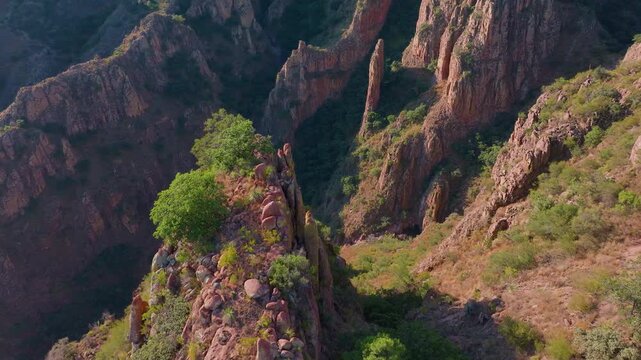 Aerial drone footage captures breathtaking rugged canyon landscape with dramatic red and brown rock spires, lush green vegetation clinging to steep cliffs, bathed in warm golden hour sunlight.