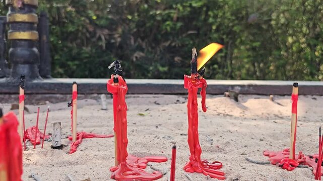 Red wax candles burning in sand at outdoor temple shrine with incense sticks and melted wax drips. Religious ritual offering with fire flame in spiritual sanctuary
