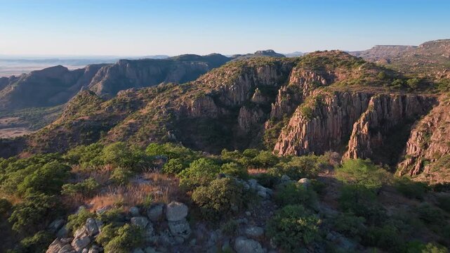 Stunning aerial footage of Sierra Madre Occidental mountains in Durango, Mexico. Dramatic red rock cliffs and green vegetation cover rugged terrain under clear blue sky during golden hour light.