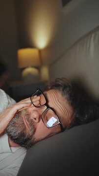 A man in eyeglasses lying in bed using smartphone late at night. 