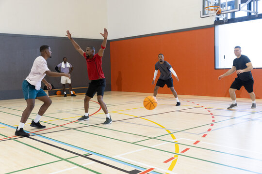Diverse male players playing pickup basketball on hardwood court, red-shirt defender raising arms