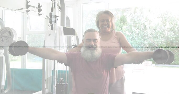Doing lateral raises bearded man holding dumbbells in home gym, woman in pink top spotting