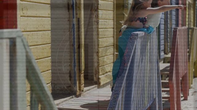 Two girls entering, leaning on railing by huts with float ring, pointing at horizon for travel