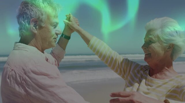 Senior lifestyle couple handholding, spinning into hug on beach with aurora drifting above for love