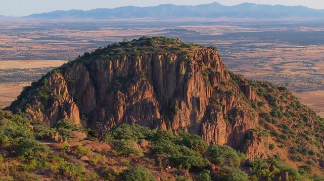 Aerial view of dramatic red rock butte with rugged cliffs and green vegetation rising from arid desert landscape in Sierra Madre Occidental near Durango, Mexico with distant mountain ranges.