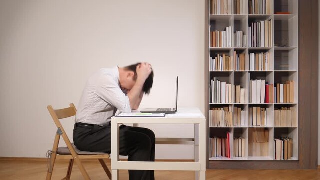 Frustrated man expressing anger and burnout, banging fists on laptop keyboard while trying to work on a white desk in a home office or study library, feeling overwhelmed by stress