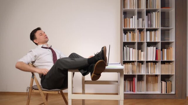 Businessman leaning back in a chair with his feet resting on the desk next to a laptop and documents, taking a break from work in a home office or study