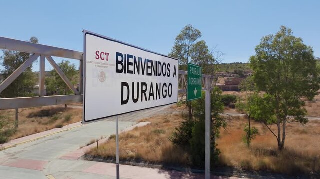 Aerial drone footage reveals Welcome to Durango Mexico highway sign with SCT government logo on metal structure over empty road with dry desert landscape and green trees under clear blue sky.