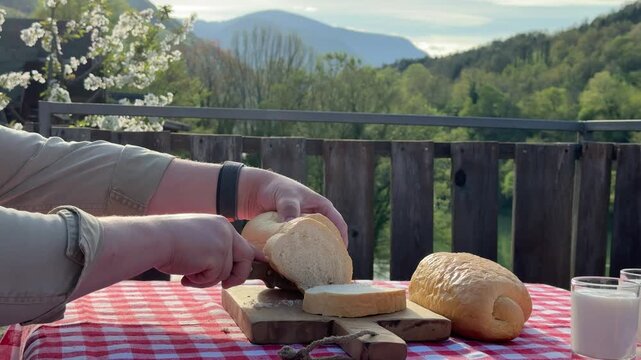 Man slicing fresh bread on wooden board at outdoor terrace with scenic mountain view in warm sunlight. Cooking outdoors and countryside lifestyle. Cozy countryside lifestyle series &mdash; outdoor cooking