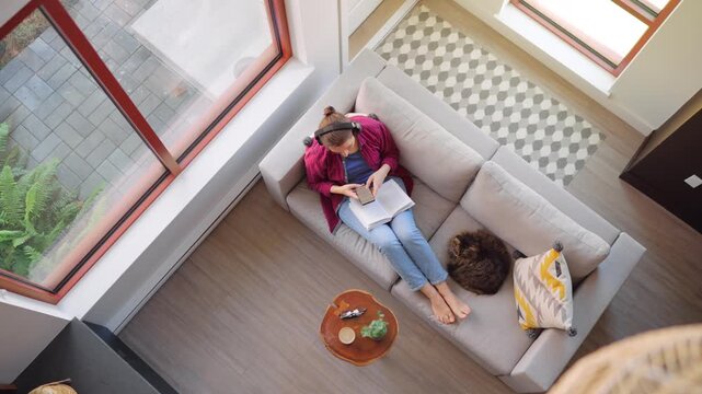 Overhead view of woman using a smartphone and reading a book on a couch at home